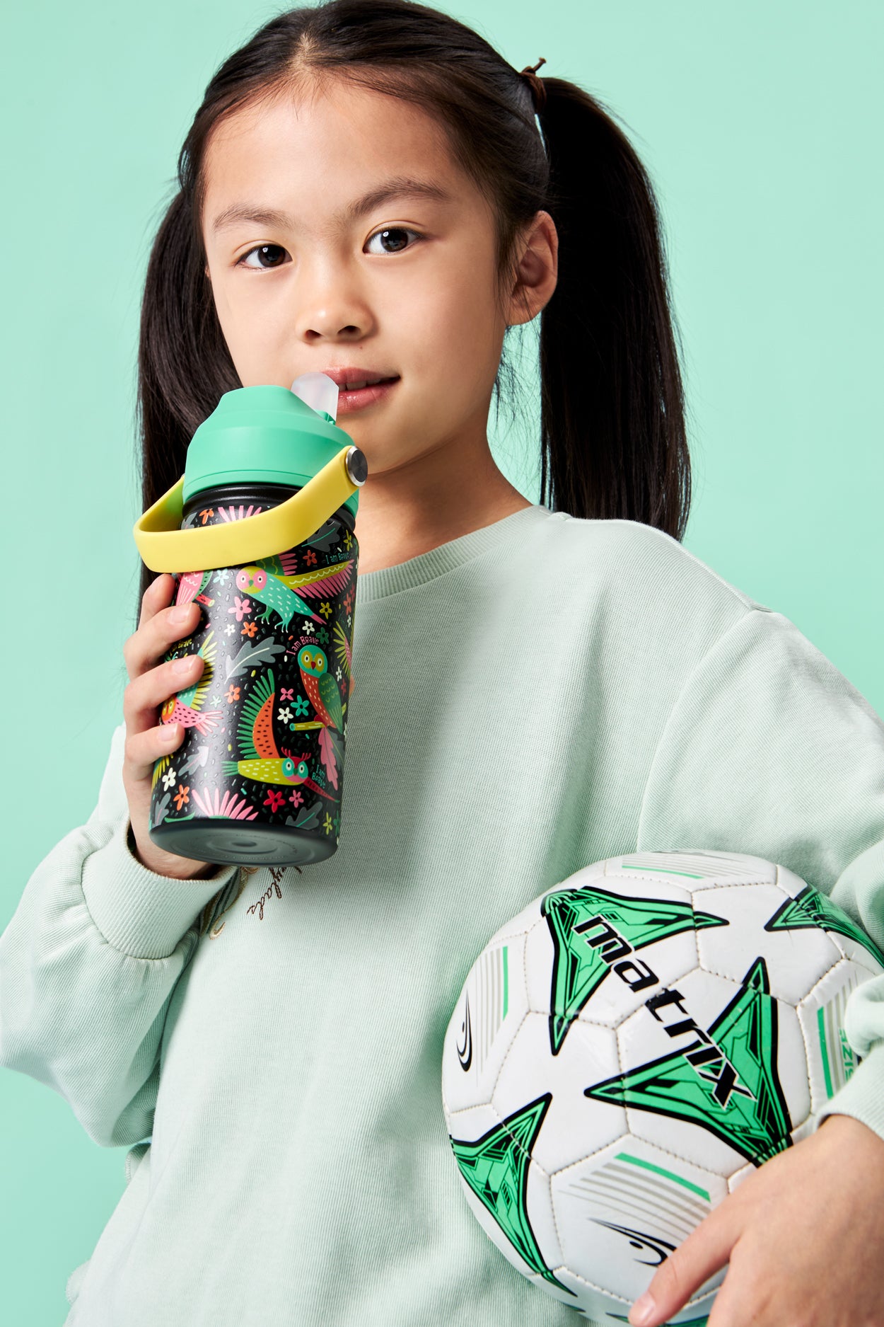 Young girl holding a sports bottle and soccer ball against a teal background